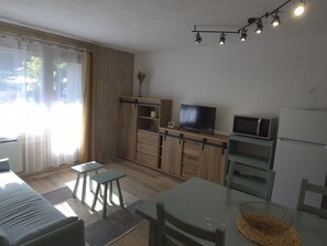 Eine moderne Küche mit Flachbildfernseher, Mikrowelle und Kühlschrank. Ein Esstisch mit Stühlen und ein kleiner Hocker stehen bereit. Der Raum verfügt über Holzschränke und ein großes Fenster mit Vorhängen.