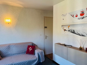 A room with a sofa, a wall-mounted light, and a wall with a ski-themed artwork. - Cozy studio, pets welcome - 2 guests (Samoëns)