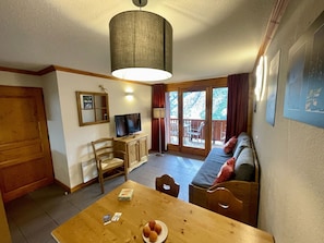Une pièce comprenant une table à manger en bois, un téléviseur posé sur un meuble en bois, un canapé et un balcon offrant une vue sur les arbres.