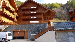 Ein mehrstöckiges Holzgebäude mit Balkonen, die mit Etage 1 bis Etage 4 beschriftet sind. Vor dem Gebäude ist ein weißer Van geparkt.