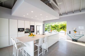 A modern kitchen with white cabinetry, a central island, and a dining area with white chairs. - Villa Magnolia Forever Luxe and Exception (St Martin)