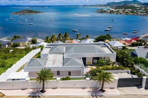 A modern building with a flat roof, surrounded by a white fence, palm trees, and a view of the sea with boats. - Villa Magnolia Forever Luxe and Exception (St Martin)