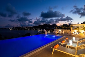 A swimming pool with lounge chairs and tables, surrounded by a mountainous landscape and a body of water at dusk. - Villa Magnolia Forever Luxe and Exception (St Martin)