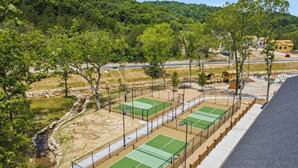 Un complexe de courts de tennis comprenant plusieurs terrains, un sentier pédestre et un paysage naturel agrémenté d’arbres et de collines en arrière-plan.