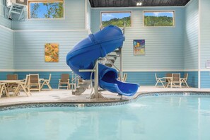 Une piscine intérieure avec un toboggan bleu, des chaises longues et une vue sur l’extérieur grâce à de grandes fenêtres.