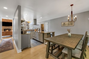 A modern kitchen with a built-in oven, a dining area with a wooden table, and a living room with a fireplace. - Woodland Dreams by Carolina Mornings | Hot Tub, Fire Pit, & Mtn Views Near AVL (Swannanoa)