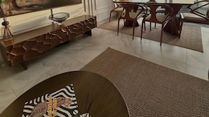 Uma sala de estar moderna com uma grande televisão de tela plana, uma área de jantar com mesa e cadeiras, e vista para o mar através de amplas janelas.