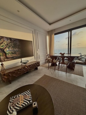 Un salon moderne doté d’un grand téléviseur à écran plat, d’un coin repas avec une table et des chaises, et offrant une vue sur la mer grâce à de grandes fenêtres.