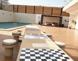 Un espace aménagé au bord de la piscine, avec un sol à carreaux, des chaises longues et un coin salon doté d’une cheminée.