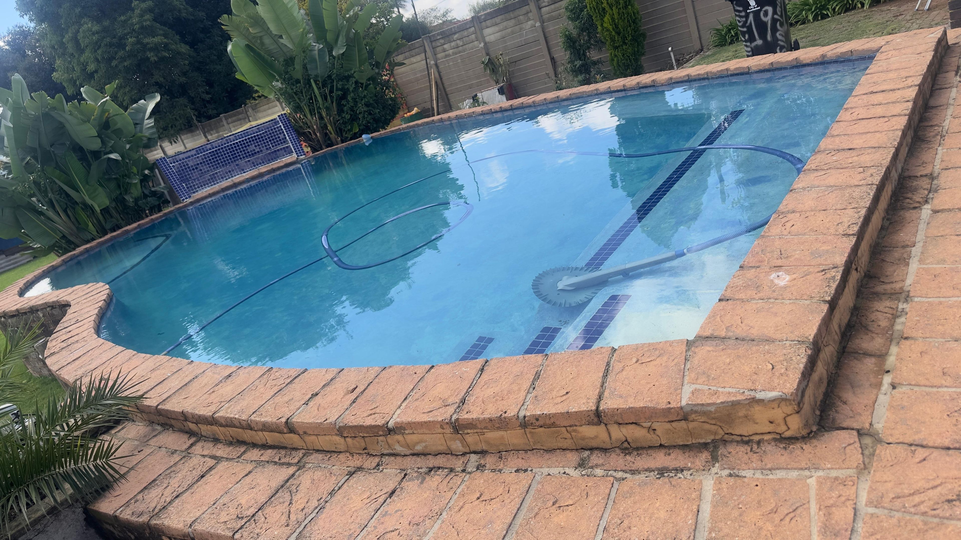Una piscina con borde de ladrillo, rodeada de vegetación y una valla.