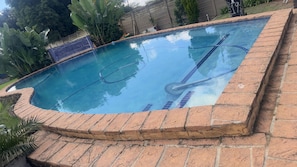 En swimmingpool med en kant av tegelstenar, omgiven av grönska och ett staket.