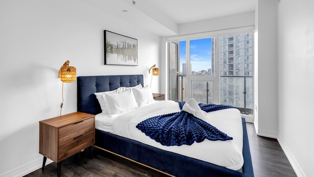 Quarto moderno com uma cama grande, mesa de cabeceira, luminária na parede e vista para a cidade através de uma janela ampla.
