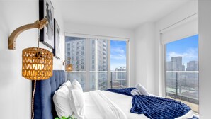 Quarto moderno com uma janela grande, colcha azul e luminária decorativa fixada na parede.