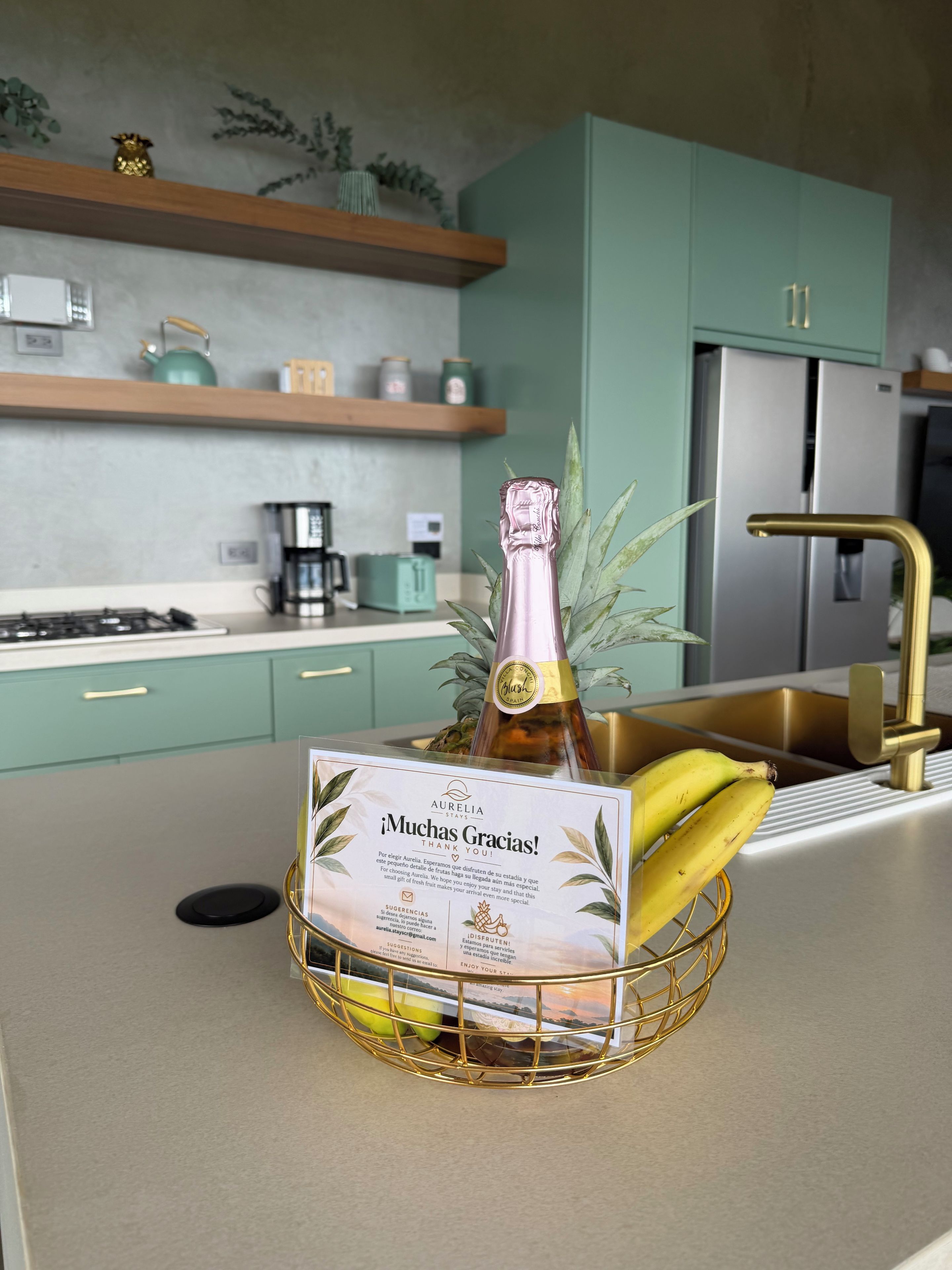Une cuisine avec des meubles de couleur turquoise, un réfrigérateur en acier inoxydable et un panier à fruits orné d’un cadre doré, contenant une bouteille de champagne et des bananes.