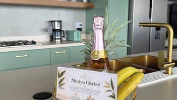 Une cuisine avec des meubles de couleur turquoise, un réfrigérateur en acier inoxydable et un panier à fruits orné d’un cadre doré, contenant une bouteille de champagne et des bananes.