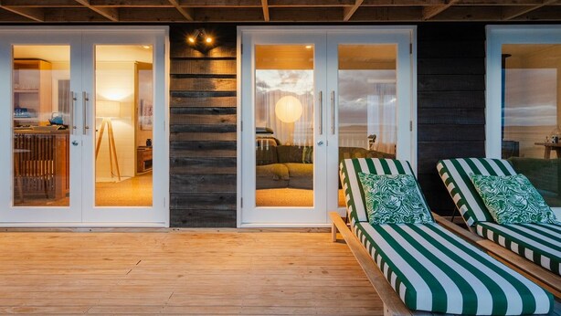 A wooden deck with a glass door entrance leading to a room with a bed and a lamp.