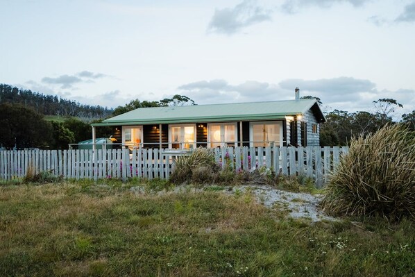 A cabin with a green roof, surrounded by a wooden fence, set in a grassy area with trees in the background.
