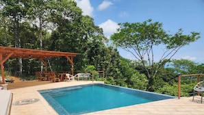 Piscina retangular com deck ladrilhado, rodeada por árvores e uma pérgula com cadeiras.