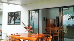 Uma área moderna para refeições ao ar livre, com mesa e cadeiras de madeira, um vaso de flores e vista do interior através de portas de vidro.