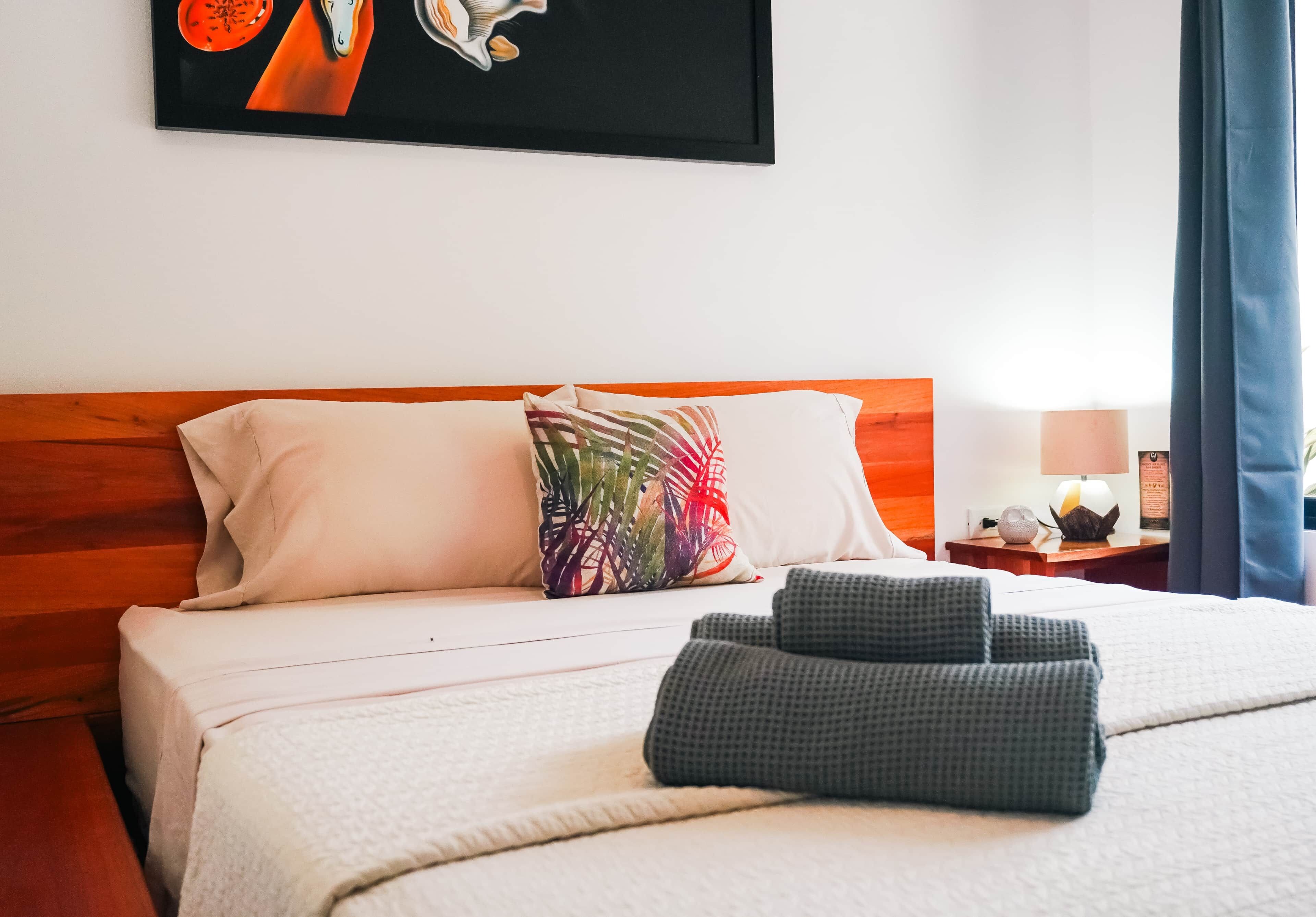 A hotel room with a bed, pillows, a blanket, a nightstand with a lamp, and a painting on the wall.