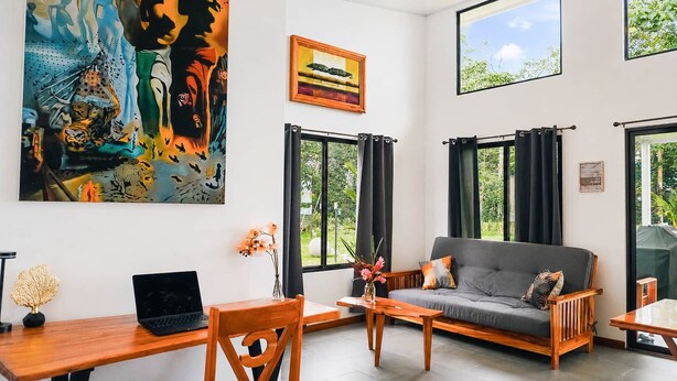 Uma sala de estar moderna com sofá, mesa de centro e escrivaninha com laptop. Há janelas amplas com cortinas, um quadro na parede e vista para o verde lá fora.