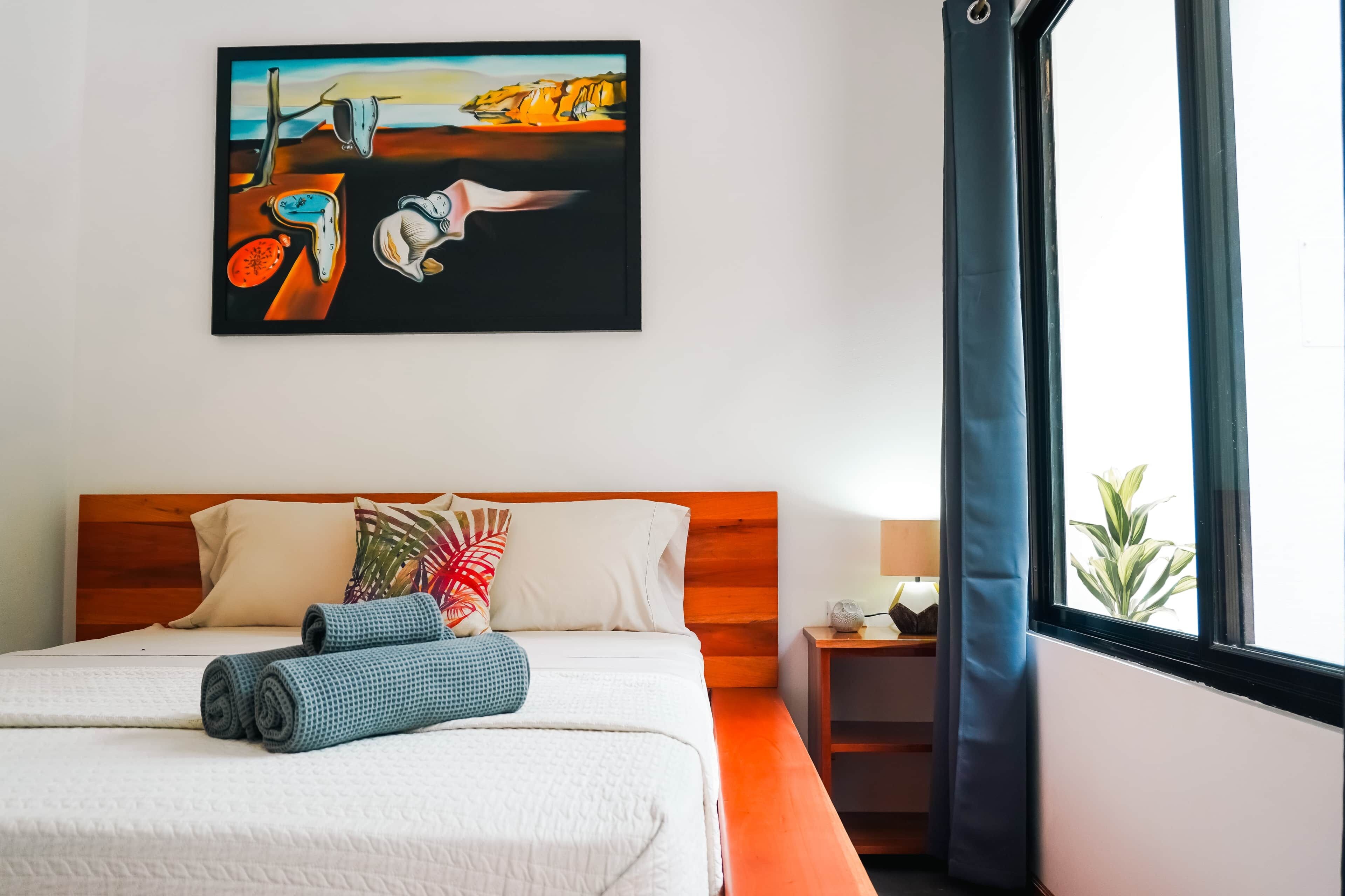 A hotel room with a bed, pillows, a painting, a nightstand, and a window with curtains.