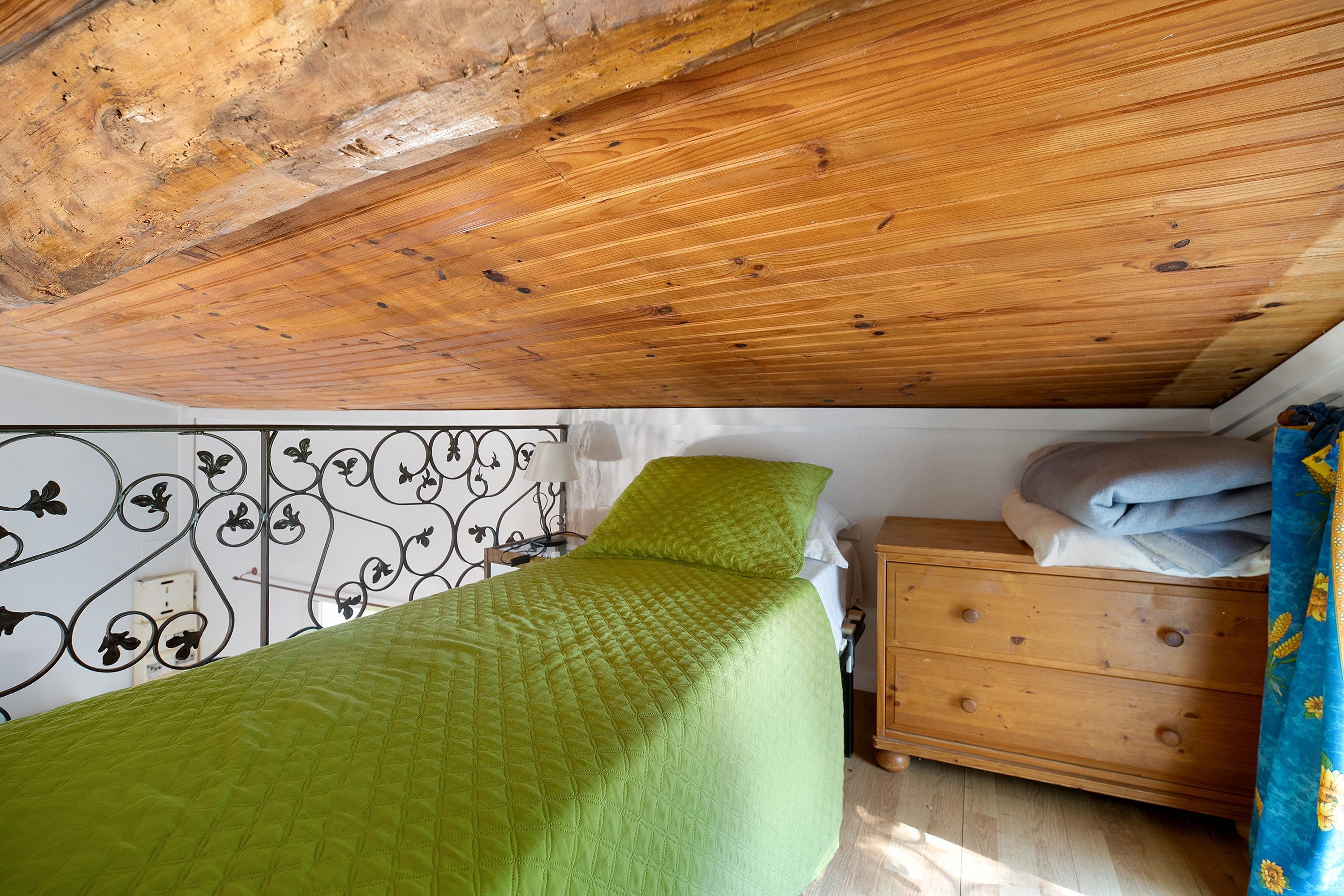 A bedroom with a wooden ceiling, a bed with a green quilt, a dresser, and a decorative metal headboard.