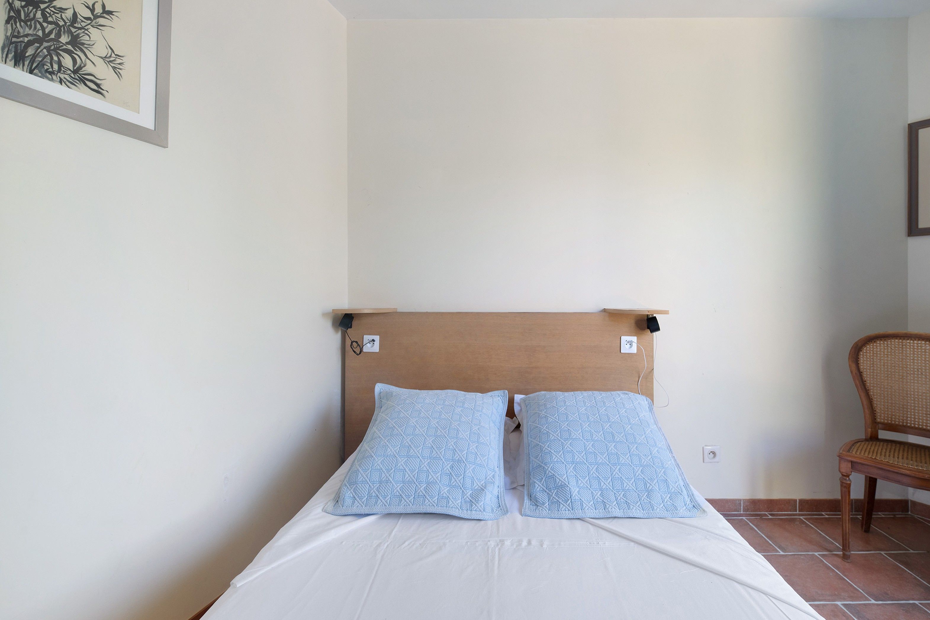 Un lit simple avec une tête de lit en bois, deux oreillers assortis, un matelas blanc et une chaise en osier dans une pièce aux murs unis et au sol carrelé.
