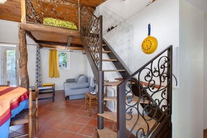 A rustic interior with a wooden staircase, a dining area with a table and chairs, a sofa, and a yellow clock on the wall.