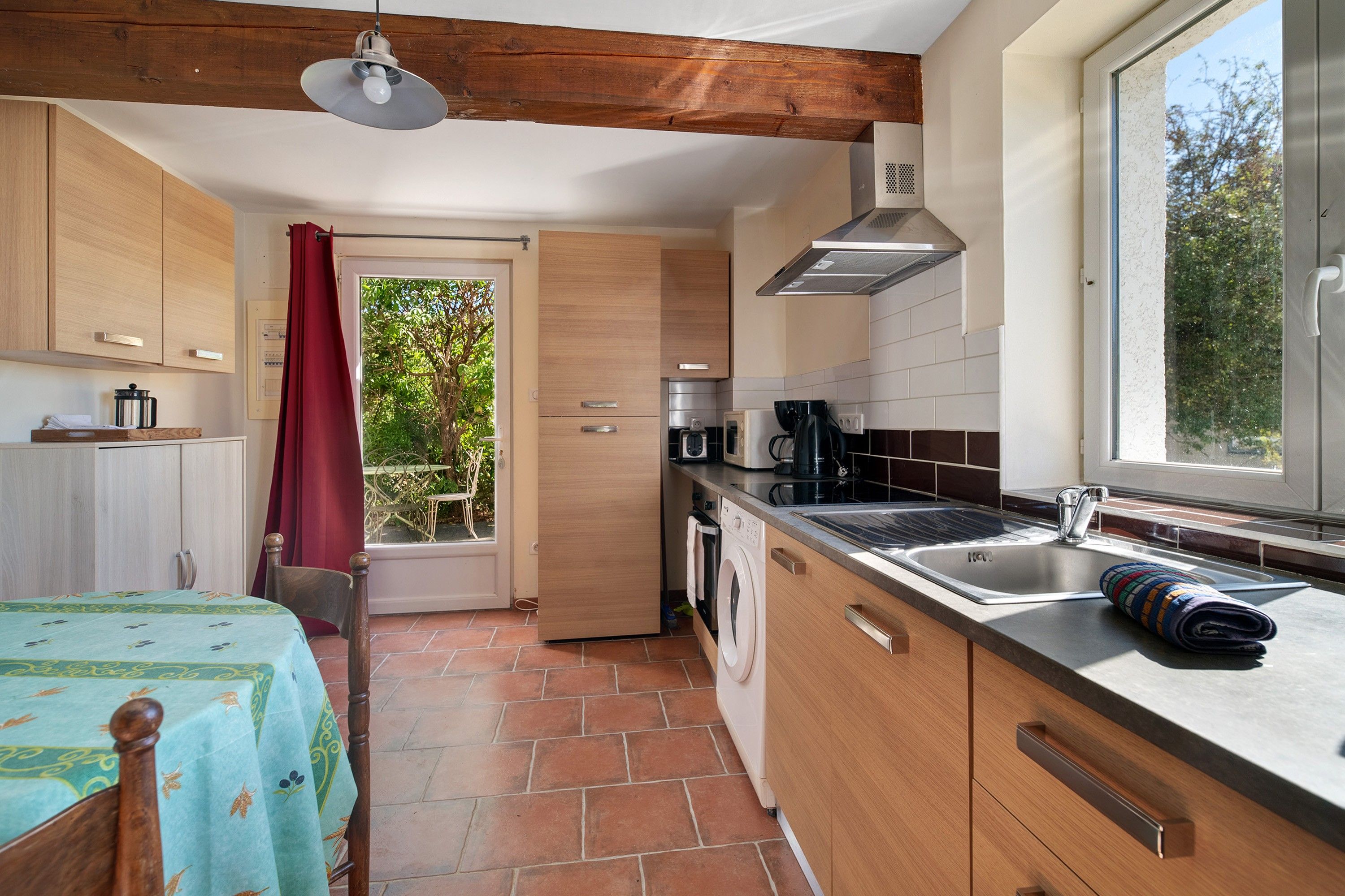 A compact kitchen with wooden cabinets, a built-in washing machine, and a window with a view of greenery.