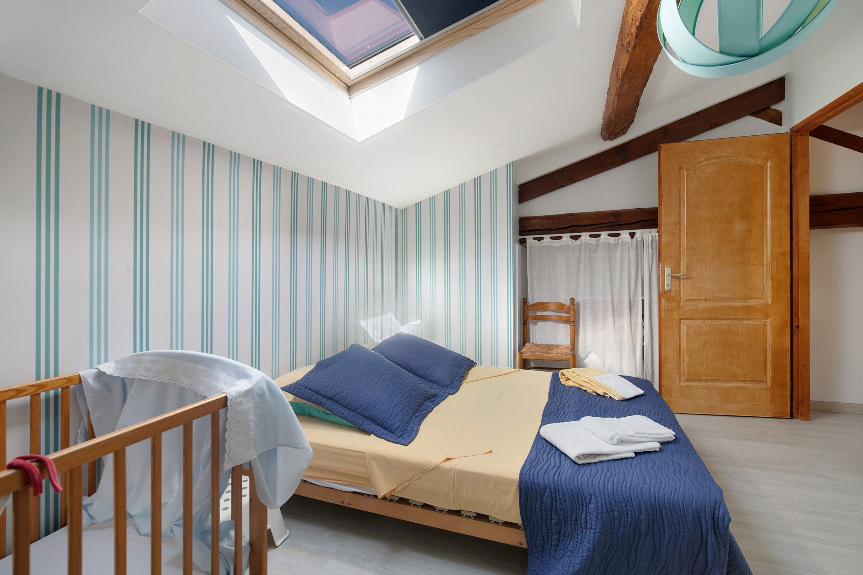 Un dormitorio con tragaluces, una cama con ropa de cama azul, una cuna de madera y una puerta.