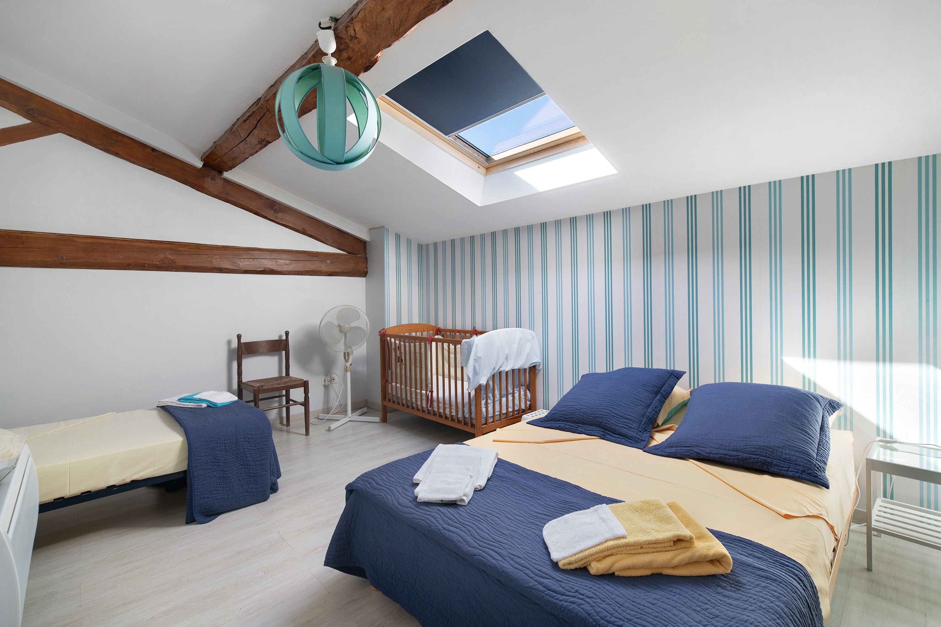 A bedroom with a skylight, a crib, a bed with blue bedding, a chair, and a fan.