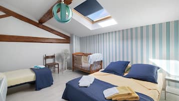 A bedroom with a skylight, a crib, a bed with blue bedding, a chair, and a fan.