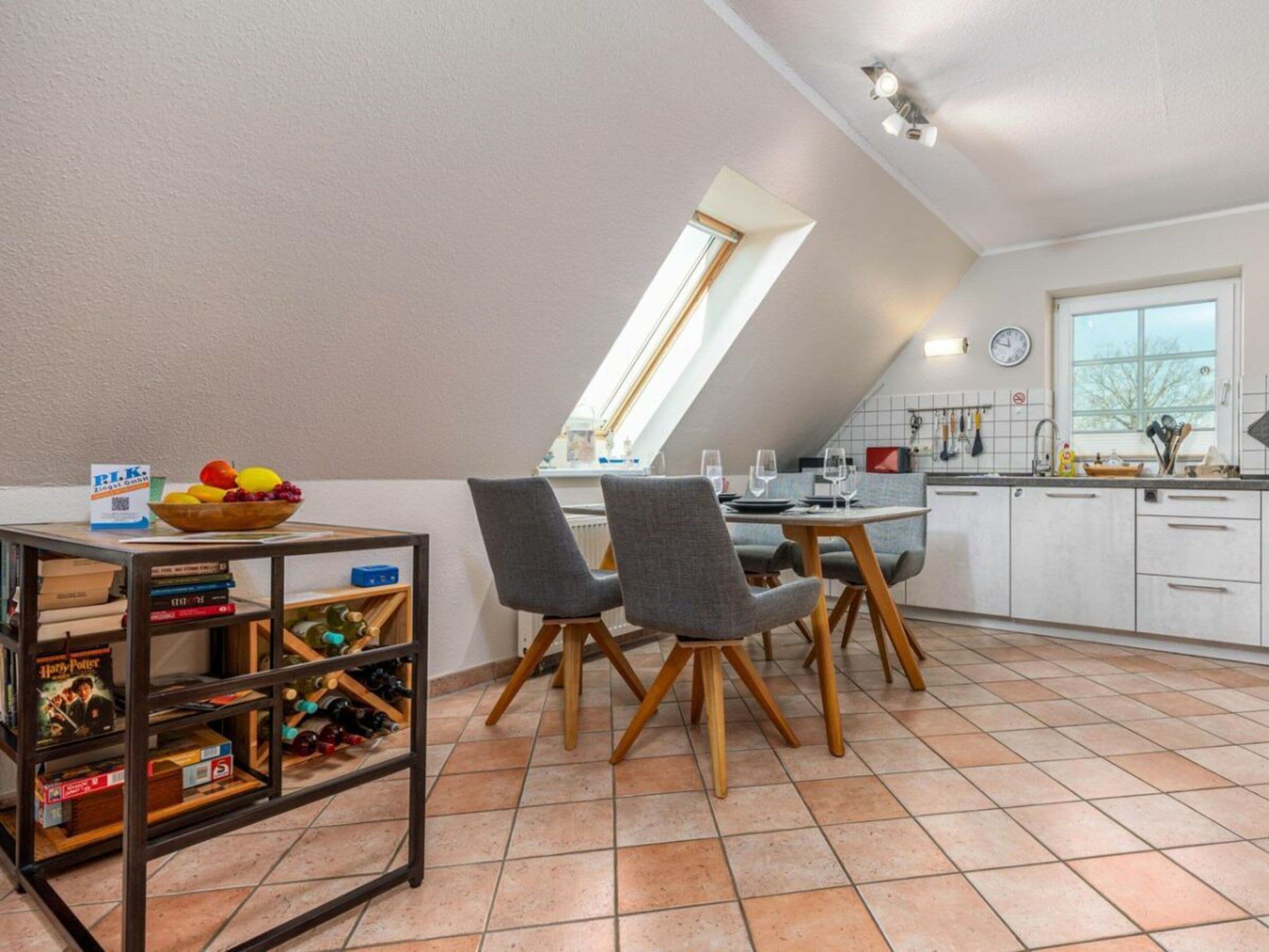A dining area with a table set for two, a bookshelf with books and a bowl of fruit, and a kitchen in the background.