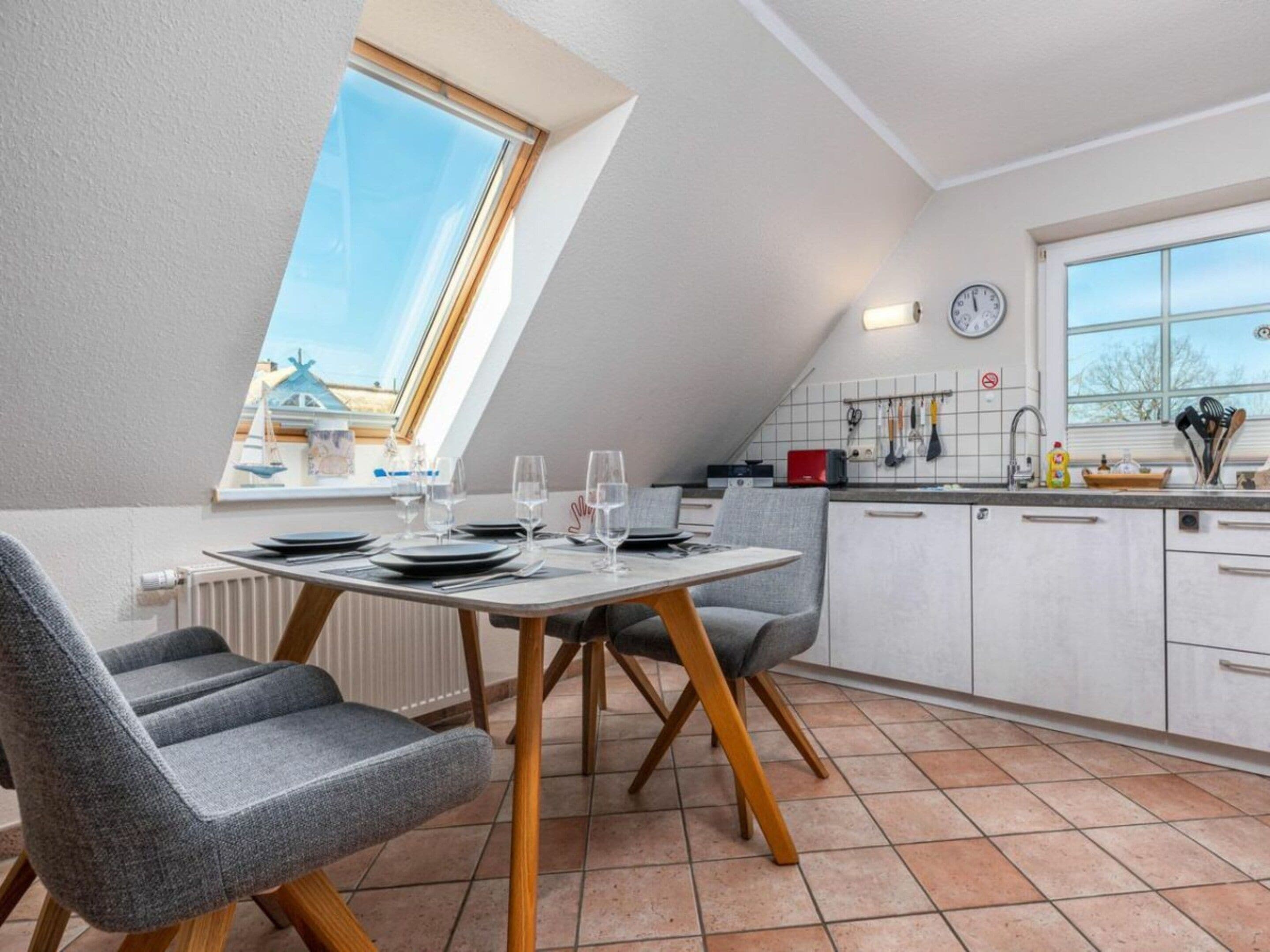 Una cocina moderna con zona de comedor, que incluye una mesa preparada para cuatro personas, un ventanal con vistas y un reloj en la pared.