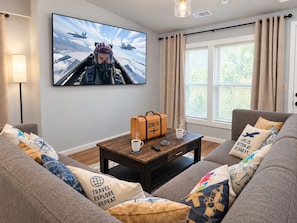 Ein Wohnzimmer mit einem großen Flachbildfernseher an der Wand, einem Couchtisch mit einem Koffer und einer Tasse sowie einer Couch mit reisebezogenen Kissen.
