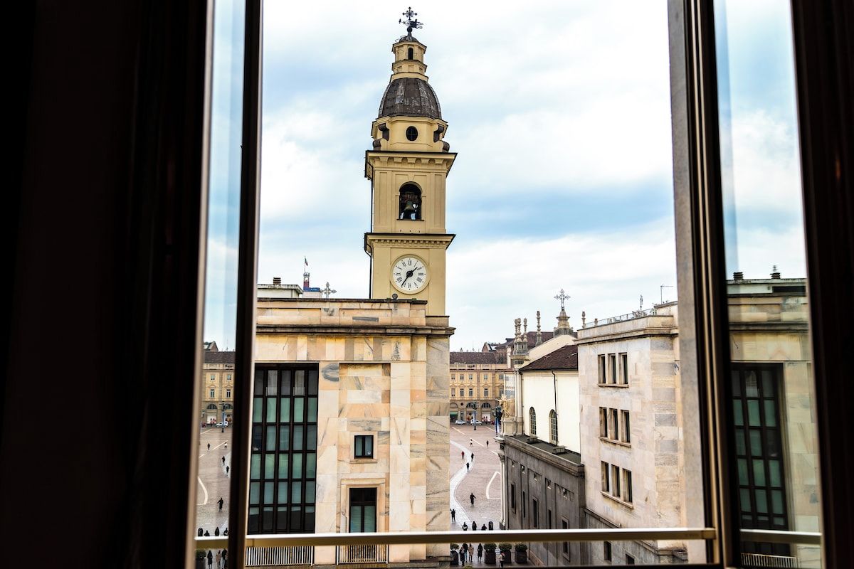 Vista de un edificio histórico con una imponente torre del reloj, a través de una ventana con marco de metal.