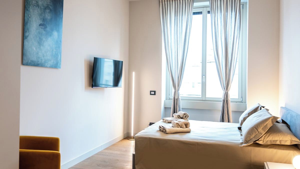 Ein Hotelzimmer mit Bett, Flachbildfernseher, Fenster mit Vorhängen und einem Bild an der Wand.