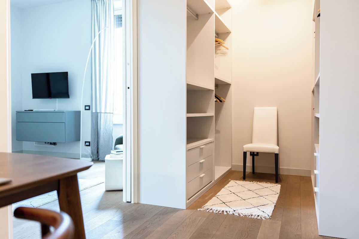 Ein modernes Zimmer mit einem Holztisch, einem weißen Stuhl, einem an der Wand montierten Fernseher und einem begehbaren Kleiderschrank mit Regalen und Schubladen.