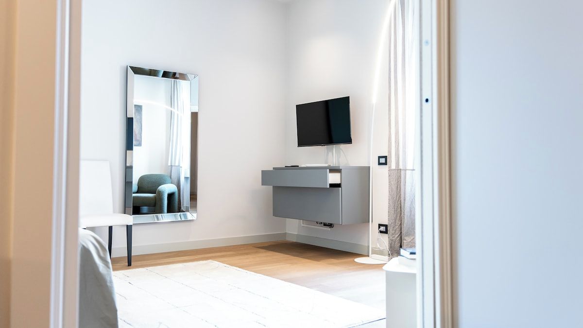 Quarto de hotel moderno com um espelho grande, televisão de tela plana, um console cinza e mesa de cabeceira branca.