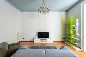 A living room with a television, a sofa, a chandelier, and a green bookshelf. - Downtown Milan FamilySuite | 6 beds w/ workspace (Milano)