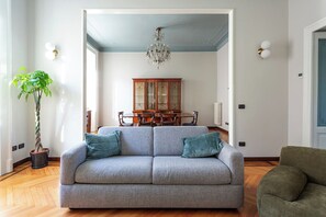 A living room with a grey sofa, a green armchair, a dining table with chairs, a chandelier, and a potted plant. - Downtown Milan FamilySuite | 6 beds w/ workspace (Milano)
