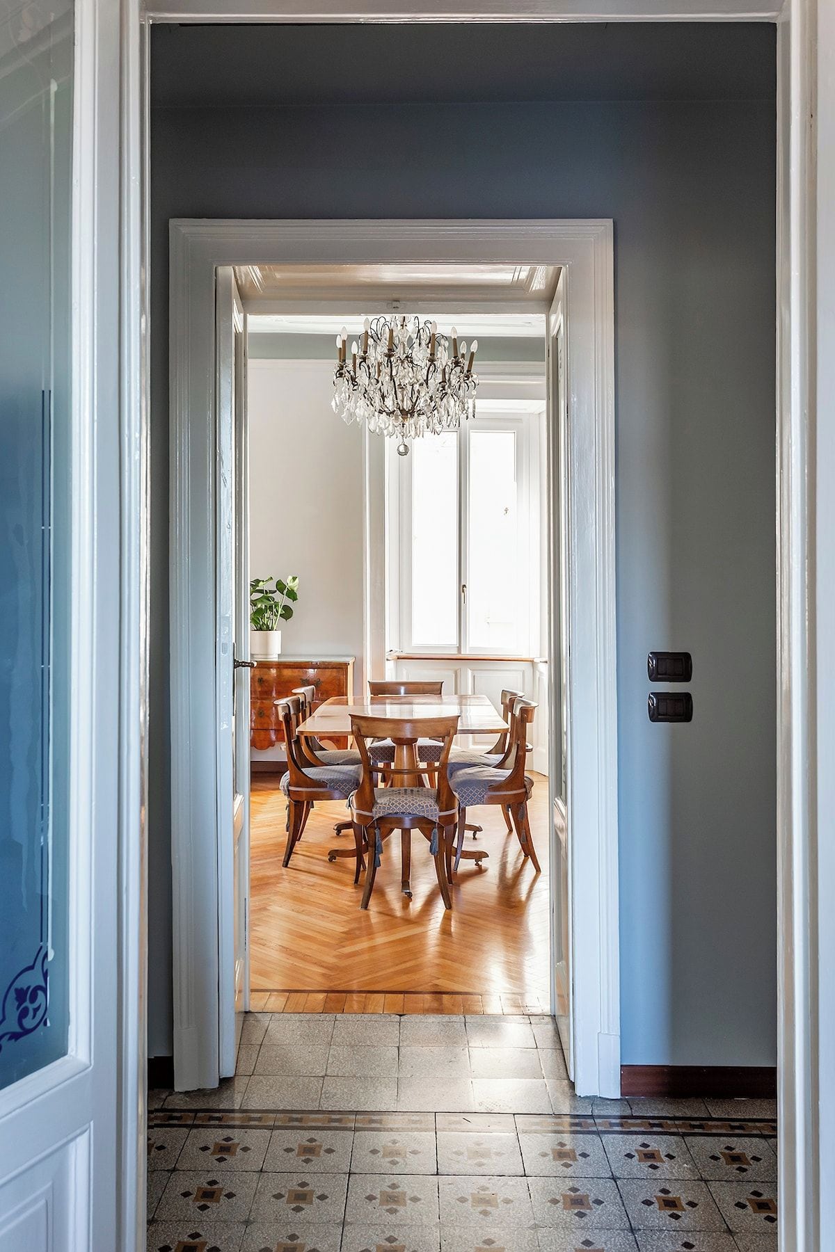 Un couloir mène à une salle à manger agrémentée d’un lustre, d’une table à manger en bois, de chaises et d’une plante en pot.