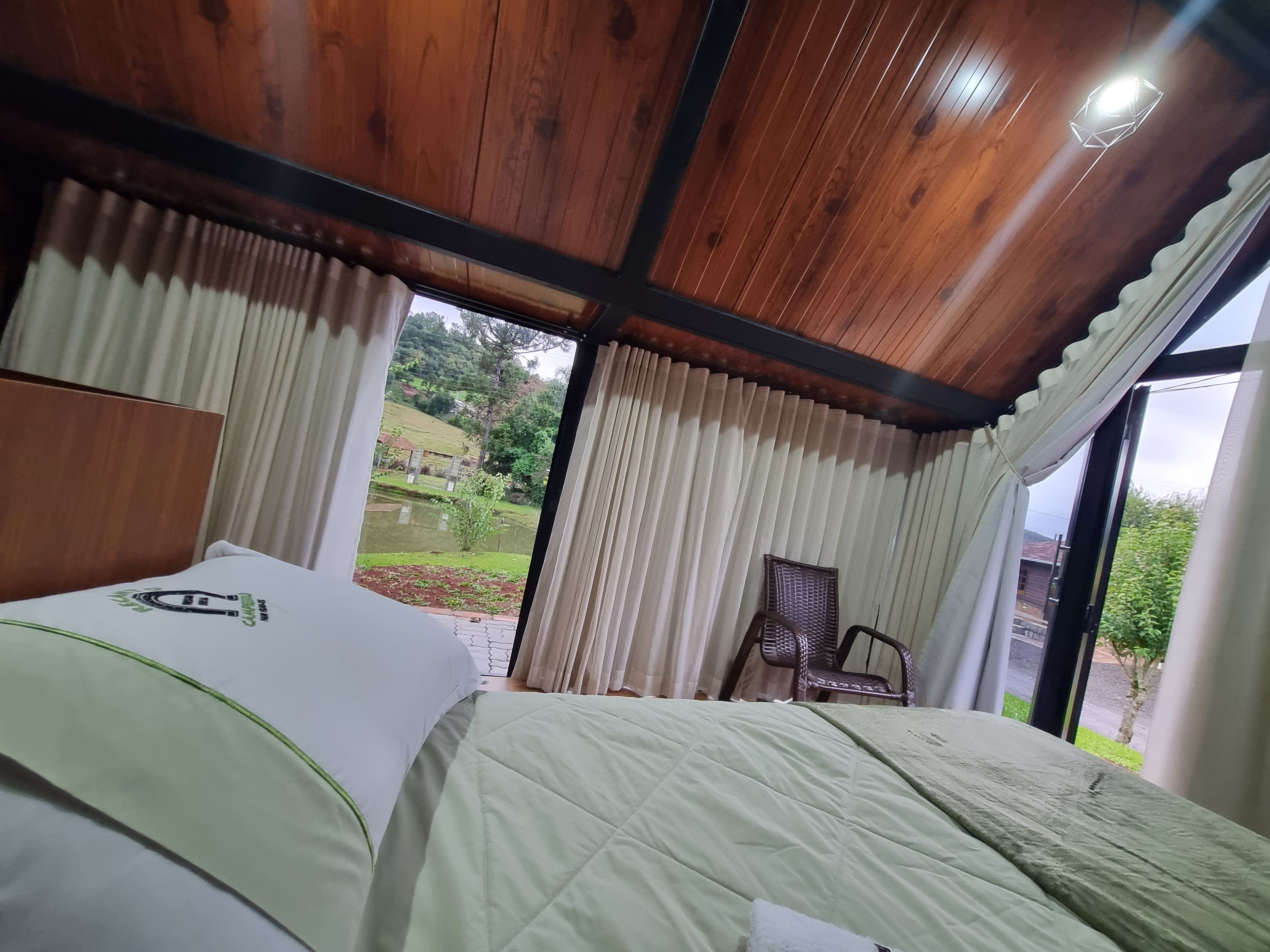 Quarto com teto de madeira, cama com colcha verde, cadeira de vime e vista para um jardim pela janela.