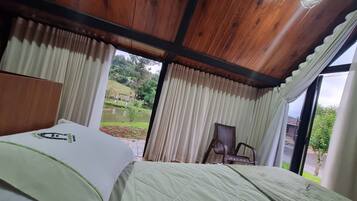 Quarto com teto de madeira, cama com colcha verde, cadeira de vime e vista para um jardim pela janela.