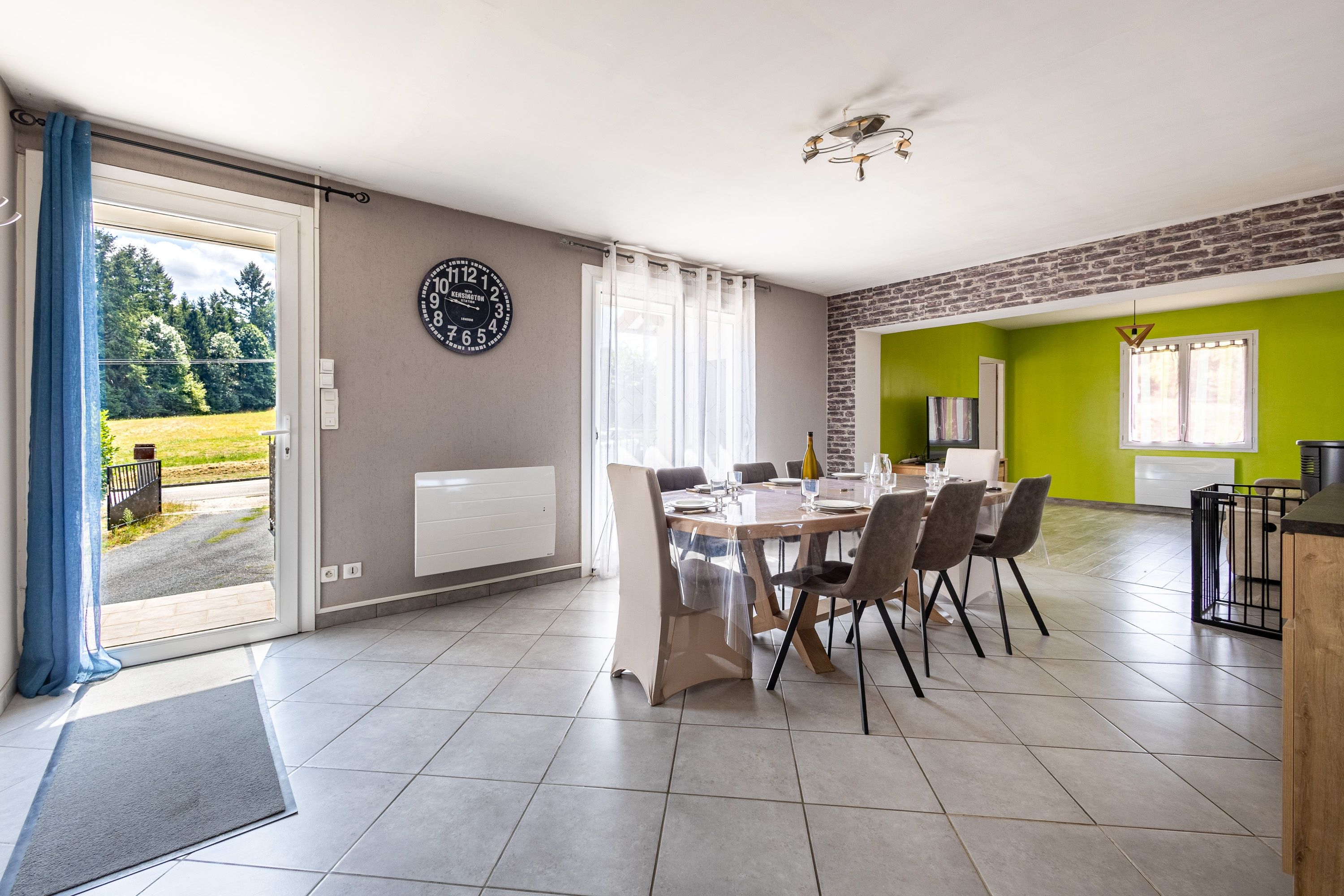 A dining area with a table set for four, a clock on the wall, and a view of a garden through the door.