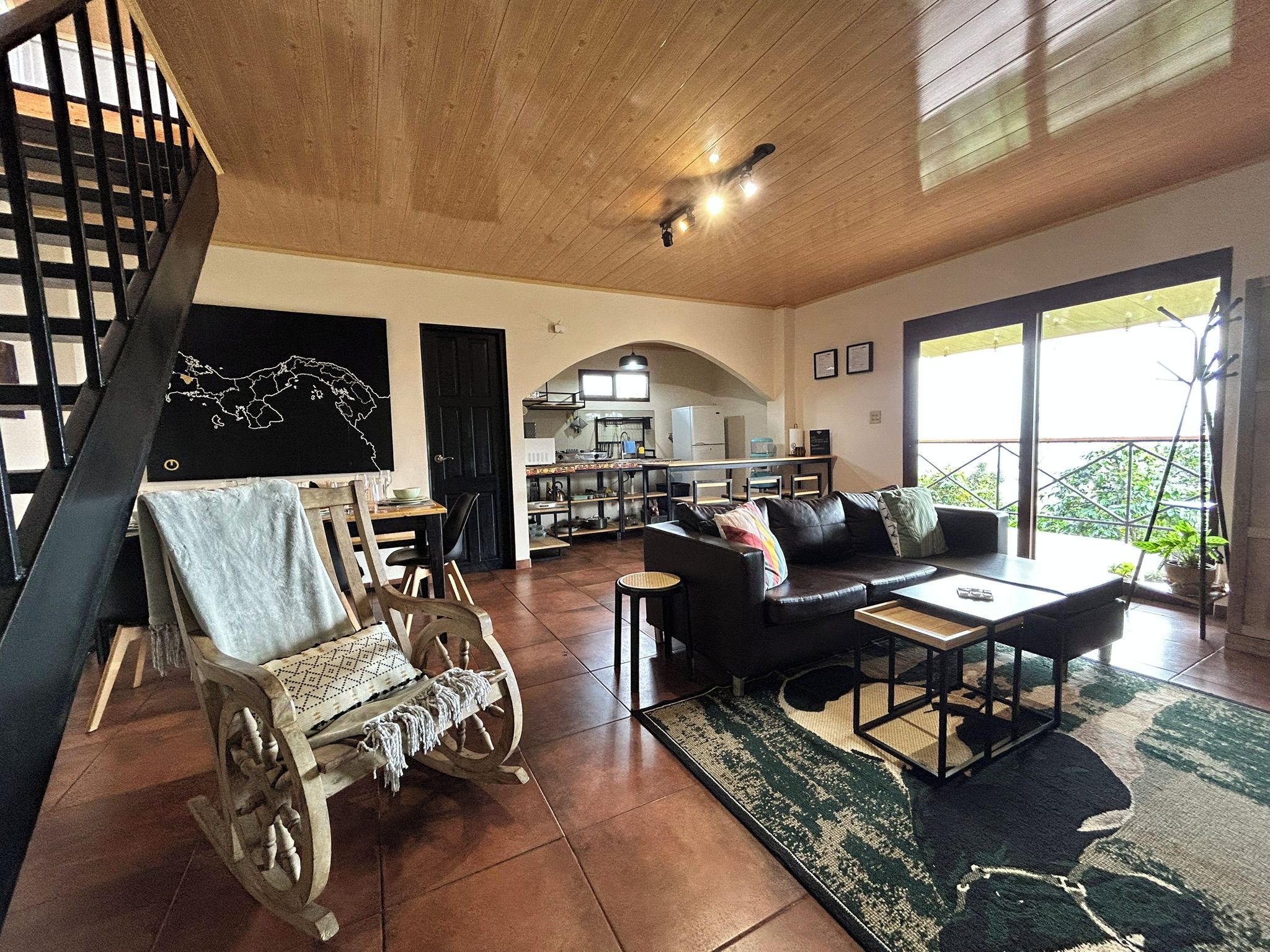A spacious living area with a wooden ceiling, a black leather sofa, a wooden rocking chair, and a dining table.