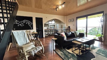 A spacious living area with a wooden ceiling, a black leather sofa, a wooden rocking chair, and a dining table.