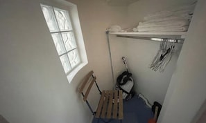 Une petite pièce avec une fenêtre, un banc en bois, un aspirateur et une étagère où sont pliées des serviettes.