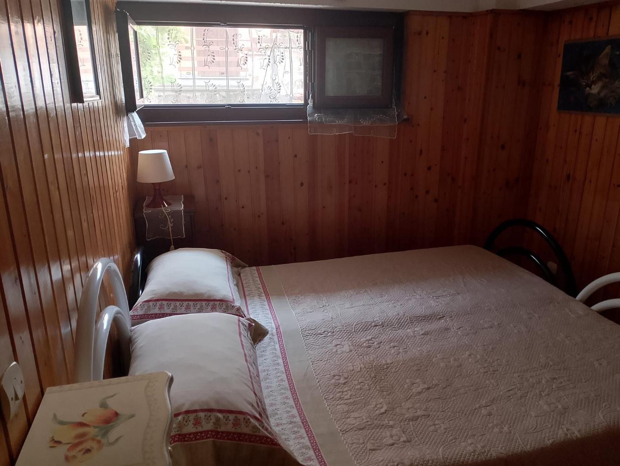 Una habitación con paredes de madera, una cama con cubrecama floral, una mesita de noche con lámpara y una ventana con vista a edificios.